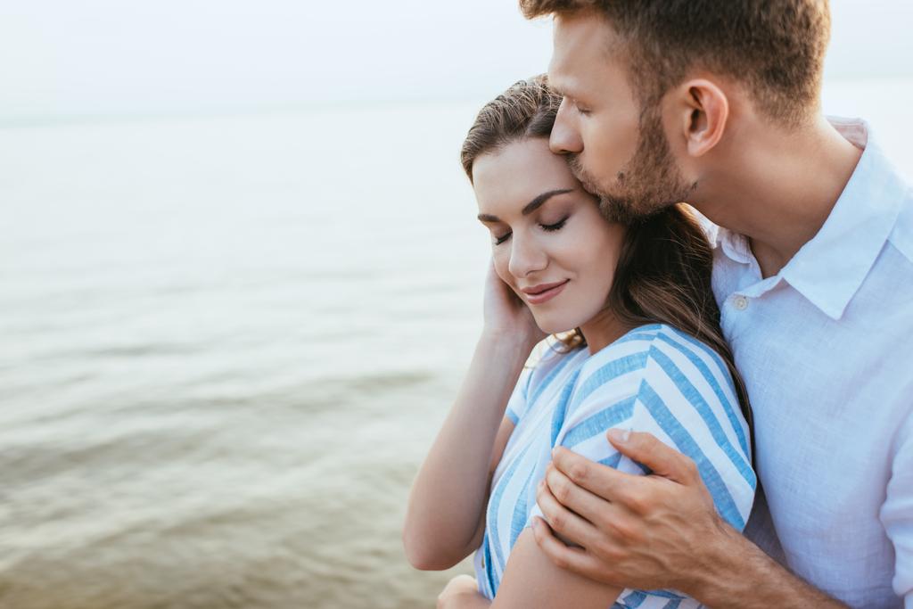 бородатый мужчина целует красивую девушку, улыбающуюся у реки - Фото, изображение
