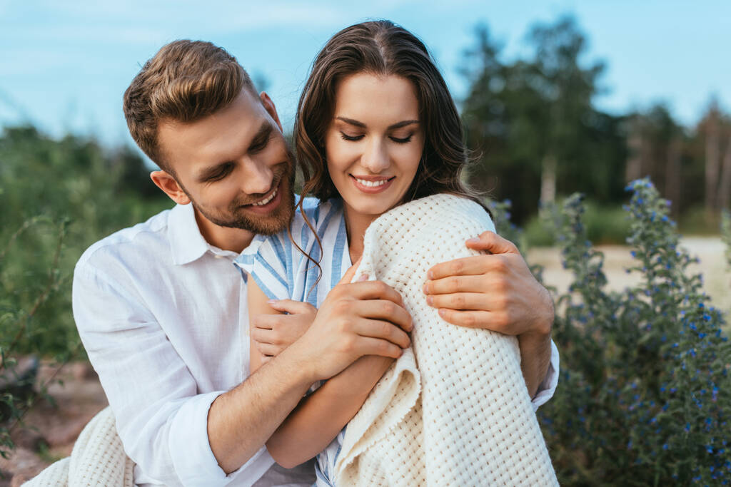 vousatý muž se usmívá a dotýká přikrývky, zatímco objímání přítelkyně venku - Fotografie, Obrázek