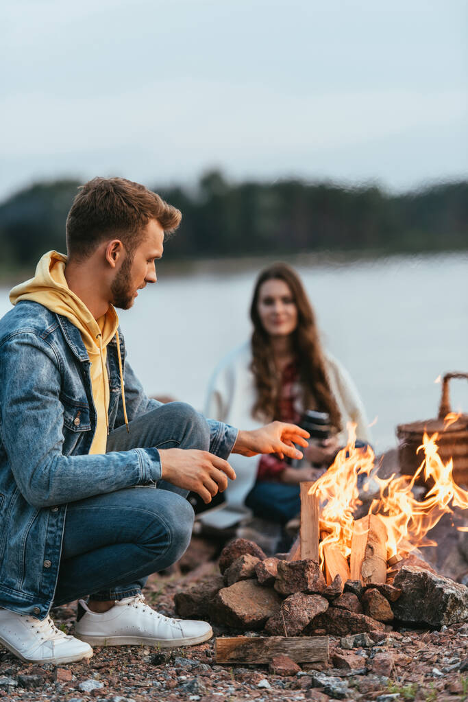 ハンサムな男の選択的な焦点は、座っているとガールフレンドの近くにたき火を燃焼を見て  - 写真・画像