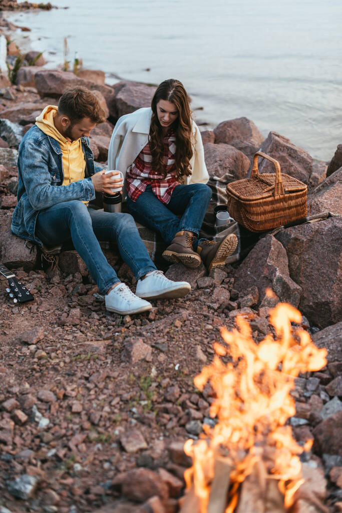 selective focus of couple sitting on stones near bonfire and lake - Photo, Image