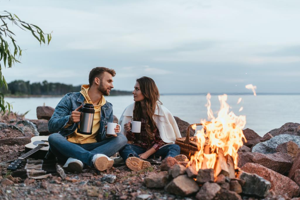 enfoque selectivo de pareja sosteniendo copas y sentado en rocas cerca de la hoguera y el lago
 - Foto, imagen