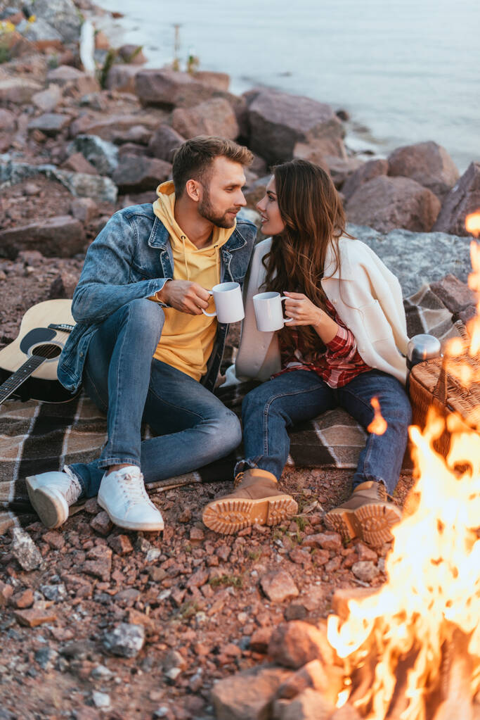 couple se regardant tout en tenant des tasses et assis sur la couverture près du feu de joie - Photo, image