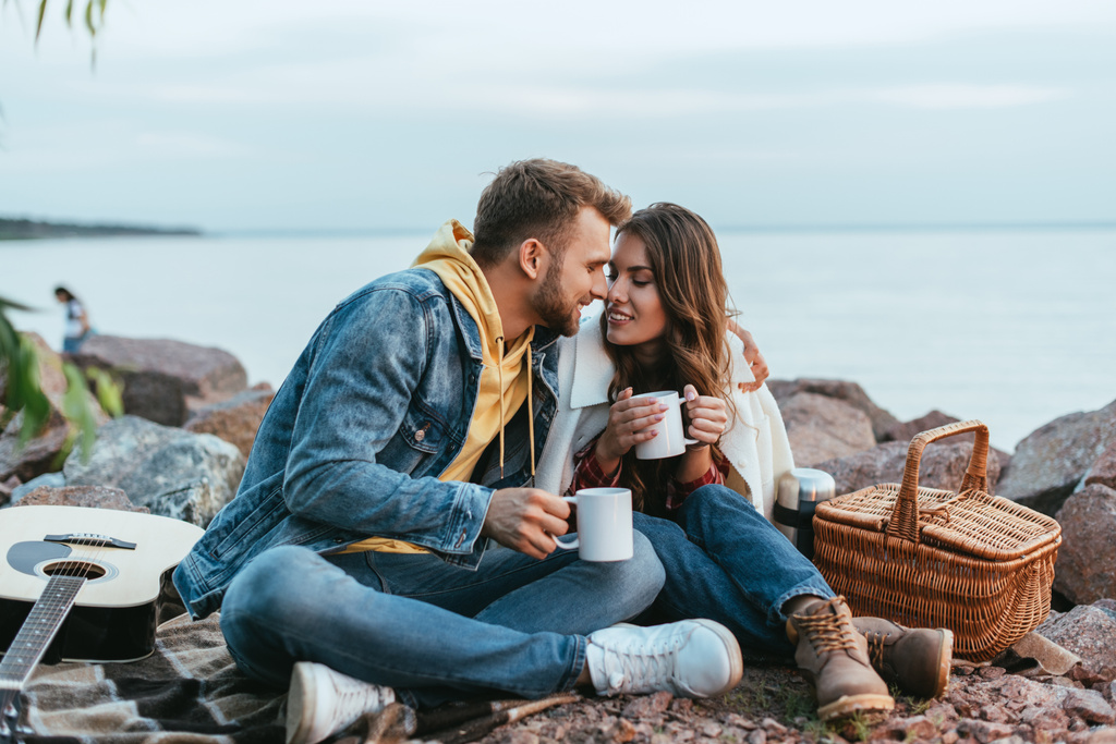 happy couple holding cups and sitting on blanket near river - Photo, Image
