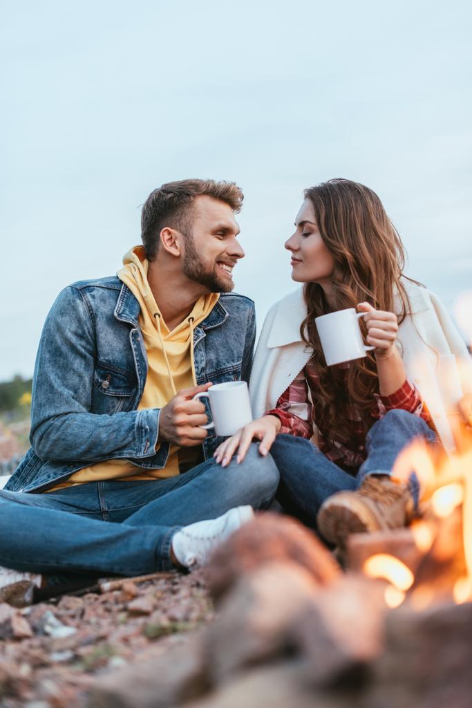 вибірковий фокус щасливого чоловіка і молодої жінки, що тримає чашки чаю біля багаття
 - Фото, зображення