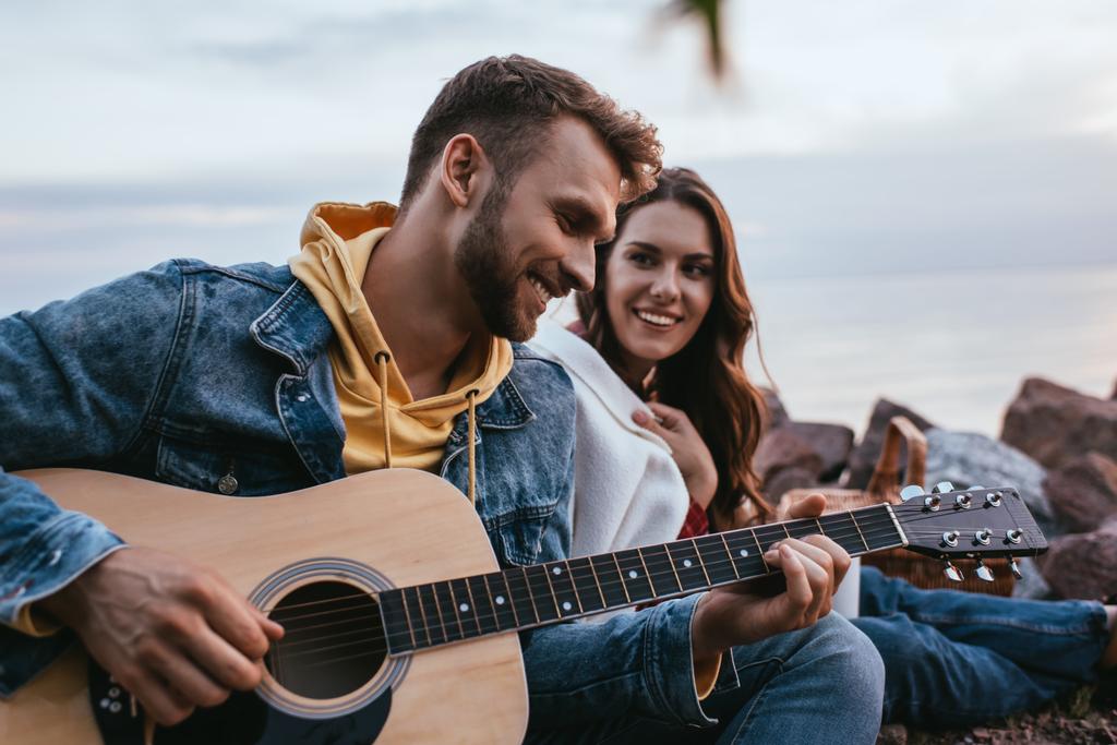 селективное внимание бородатого мужчины, играющего на акустической гитаре рядом с подругой и костром - Фото, изображение