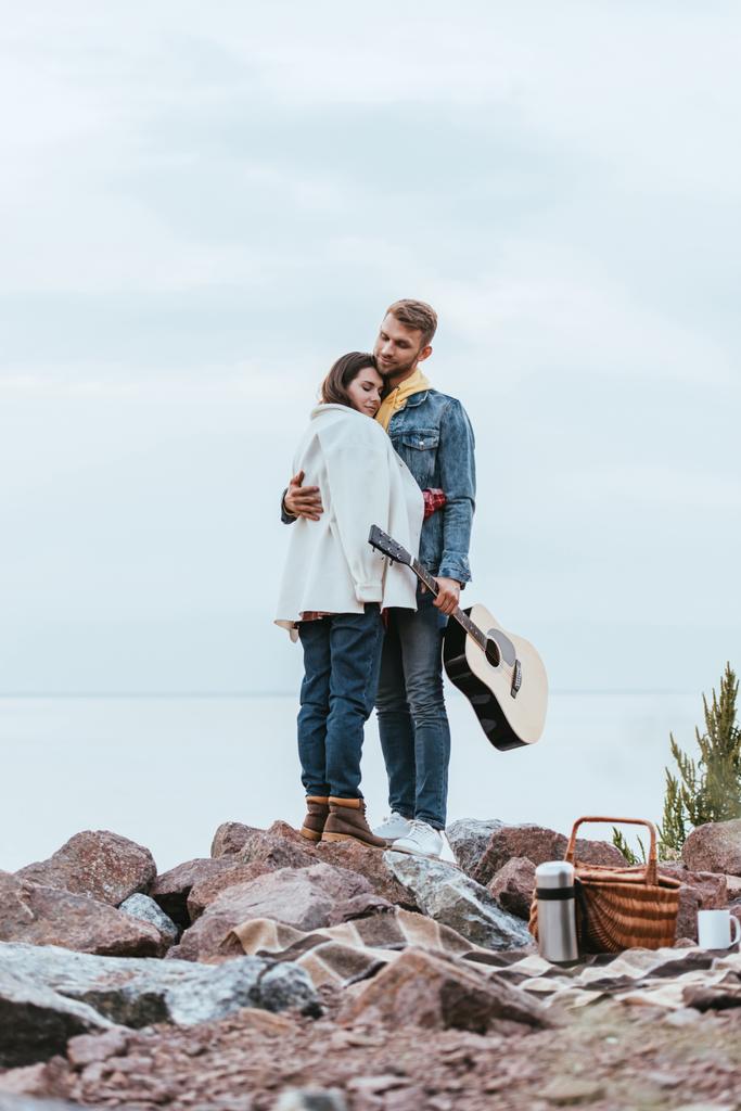 όμορφος άντρας κρατώντας ακουστική κιθάρα και αγκαλιάζει το κορίτσι ενώ στέκεται κοντά στο ποτάμι  - Φωτογραφία, εικόνα