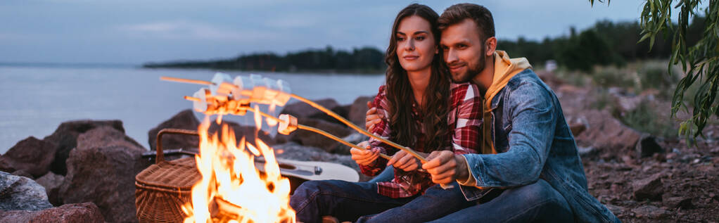 たき火の近くの棒でマシュマロを焙煎する夫婦のパノラマ写真  - 写真・画像