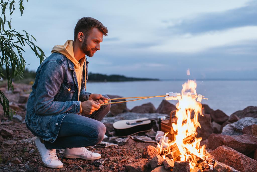 homem feliz assar marshmallows em paus perto de fogueira
  - Foto, Imagem