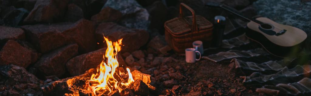 Ekose battaniye, hasır sepet, fincanlar ve akustik gitar yanında yanan şenlik ateşinin panoramik mahsulü  - Fotoğraf, Görsel
