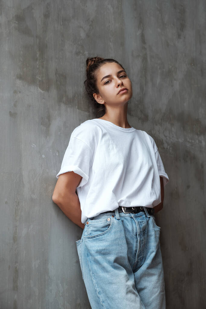 A sad pensive girl stands at the wall. Stylish young woman in blue jeans and a white T-shirt stands against the background of a gray concrete wall. - Photo, Image