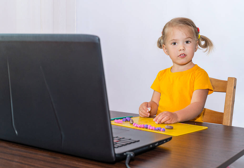 Una niña interesada mira su computadora portátil y esculpe de plastilina. La niña estudia en casa y hace los deberes escolares. Un hogar de aprendizaje a distancia
. - Foto, Imagen