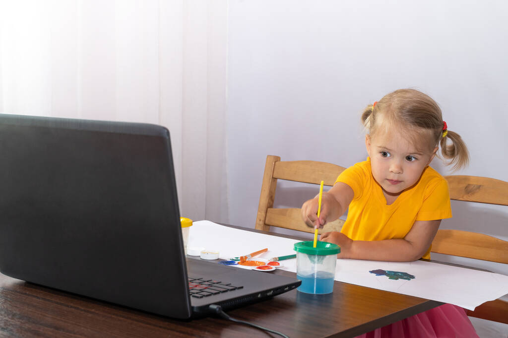 Ένα ενδιαφερόμενο κοριτσάκι κοιτάζει το λάπτοπ της και ζωγραφίζει. Online μαθήματα. - Φωτογραφία, εικόνα