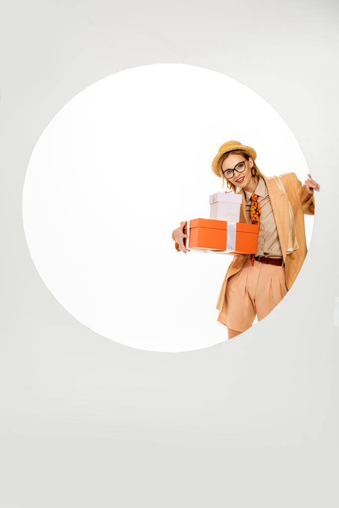 Mulher na moda sorrindo enquanto segurando presentes perto buraco redondo no fundo branco
  - Foto, Imagem