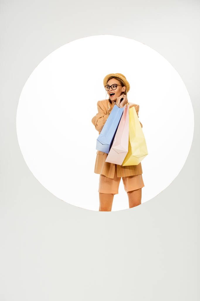 Neşeli kız beyaz arka planda, yuvarlak deliğin arkasında renkli alışveriş torbaları tutuyor.  - Fotoğraf, Görsel