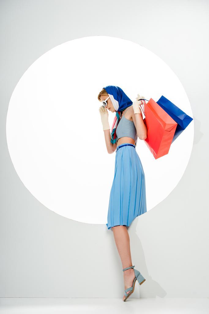 Вид сбоку на триумфальную женщину, держащую красные и синие сумки для покупок возле круглой дыры на белом фоне  - Фото, изображение