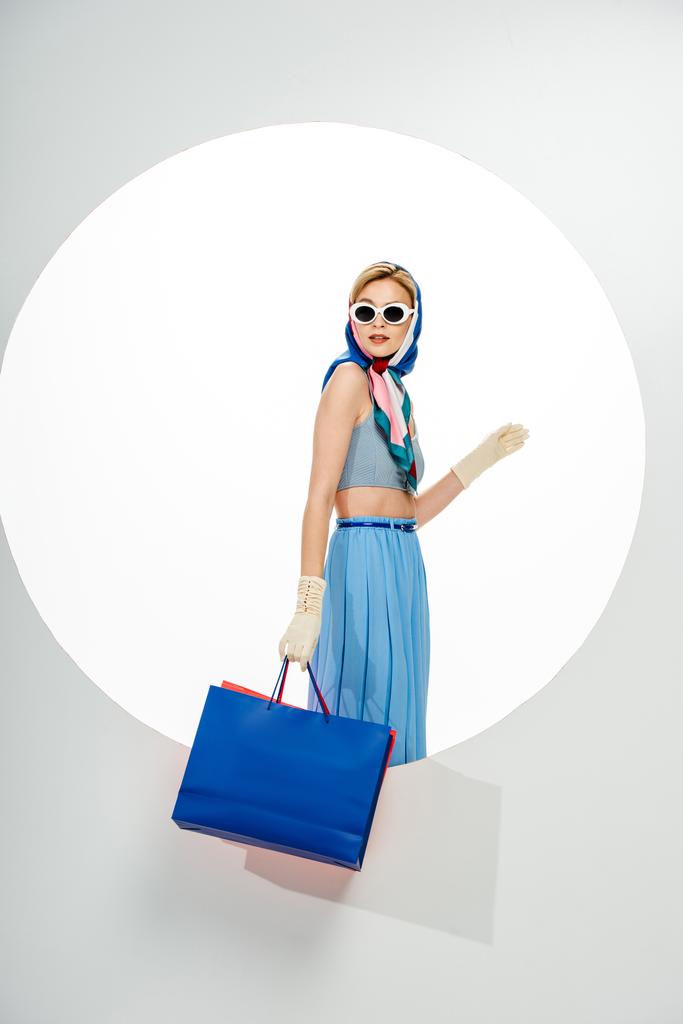 Menina na moda em lenço de cabeça e óculos de sol segurando sacos de compras perto do círculo no fundo branco
  - Foto, Imagem