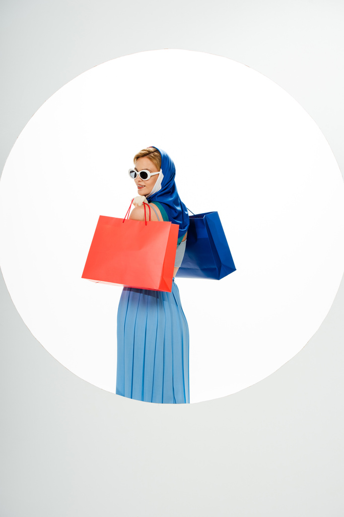 Back view of smiling woman in headscarf and sunglasses holding red and blue shopping bags behind circle on white background  - Photo, Image