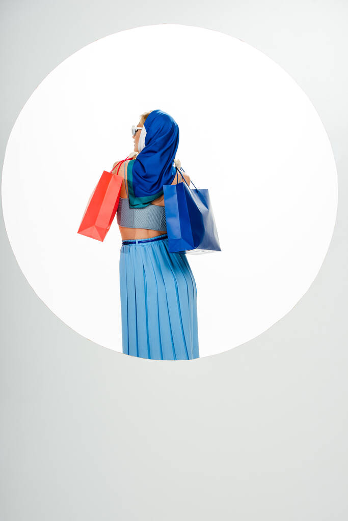 Powrót widok stylowej kobiety w chustce trzymającej czerwone i niebieskie torby na zakupy w pobliżu koła na białym tle  - Zdjęcie, obraz