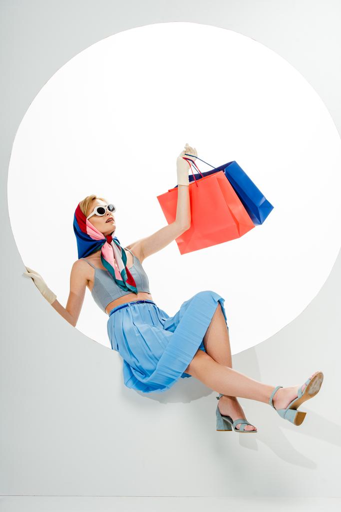 Güneş gözlüklü modaya uygun bir kadın beyaz arka planda mavi ve kırmızı alışveriş torbaları arasında poz veriyor.  - Fotoğraf, Görsel
