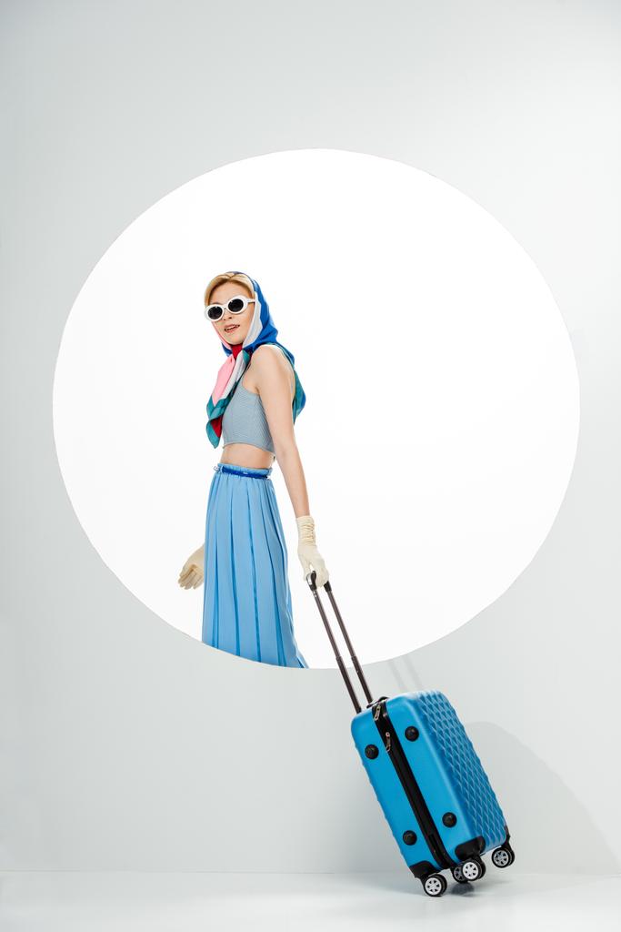 Stilvolle Frau mit Sonnenbrille und blauem Koffer in Kreisnähe auf weißem Hintergrund  - Foto, Bild
