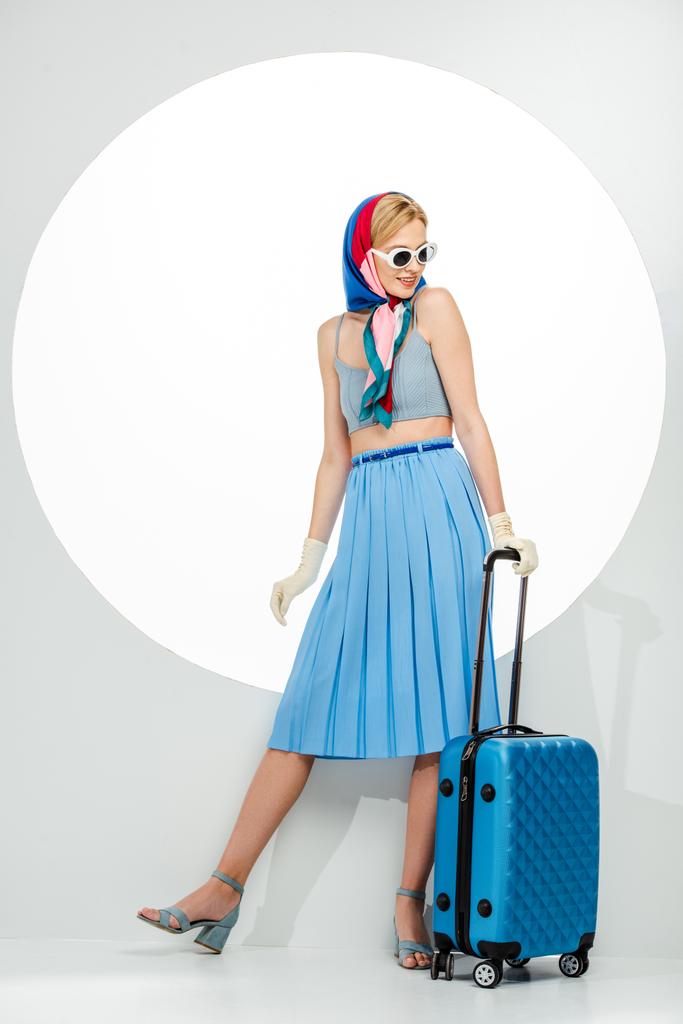 Uśmiechnięta stylowa kobieta w okularach przeciwsłonecznych trzymająca niebieską walizkę na białym tle - Zdjęcie, obraz