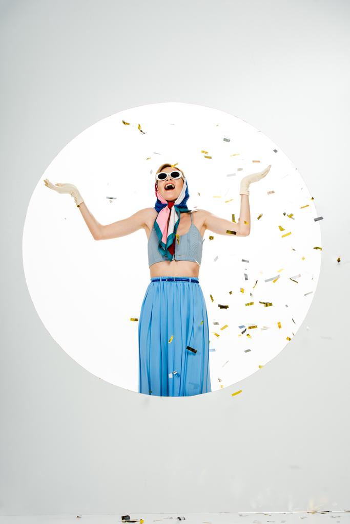 Geschokt stijlvolle vrouw staan onder vallende confetti in de buurt cirkel op witte achtergrond - Foto, afbeelding