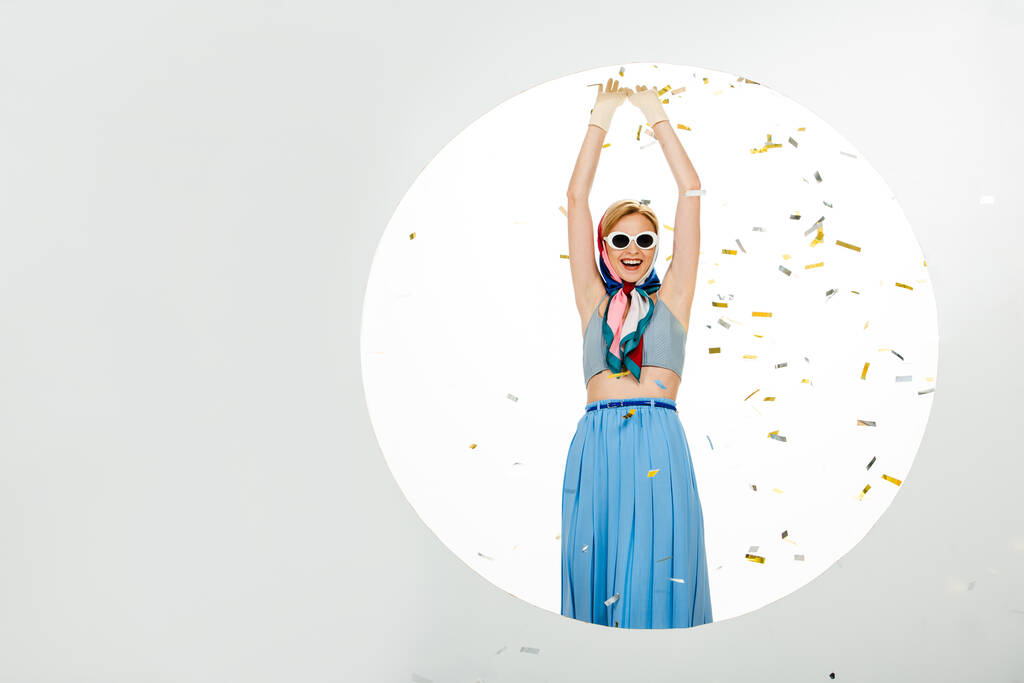Позитивная девушка в солнечных очках, стоящая возле круга под падающими конфетти на белом фоне - Фото, изображение