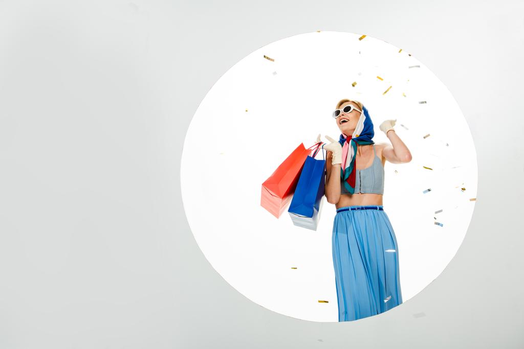 Veselý stylový dívka drží nákupní tašky pod pádu konfety v blízkosti kruhu na bílém pozadí - Fotografie, Obrázek