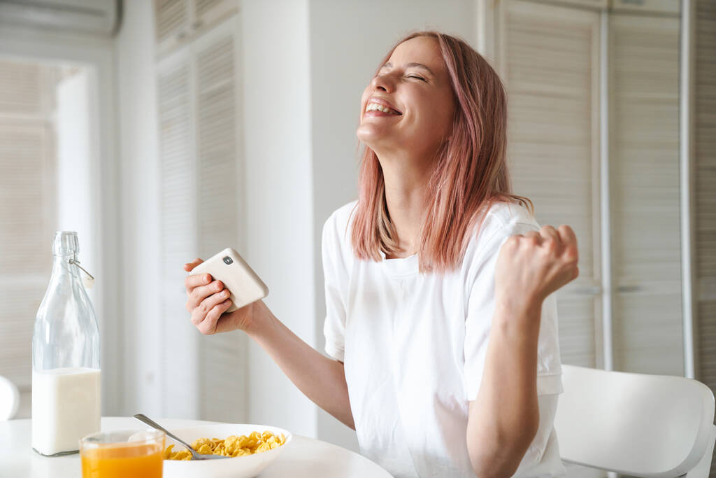 Foto de una joven mujer emocionada haciendo gesto ganador mientras juega juego en línea en el teléfono celular en la cocina blanca
 - Foto, imagen