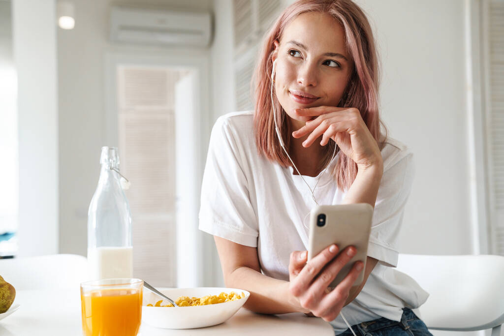 Zdjęcie miłej zadowolonej kobiety korzystającej z telefonu komórkowego i słuchawek podczas śniadania w białej kuchni - Zdjęcie, obraz