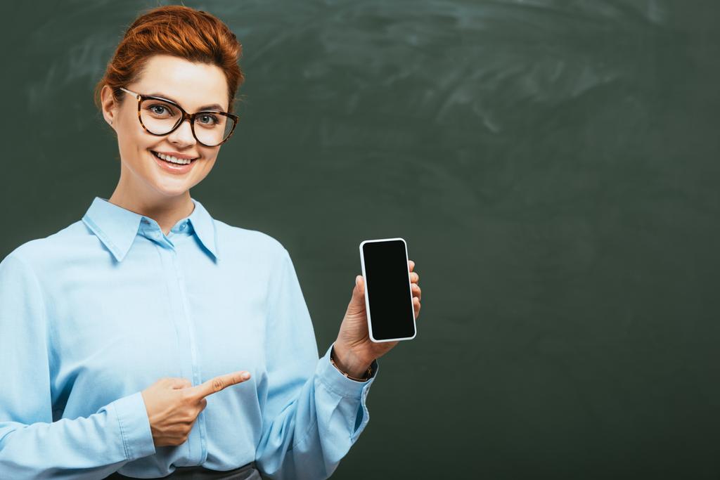 professeur heureux dans les lunettes pointant vers smartphone avec écran blanc près du tableau noir  - Photo, image