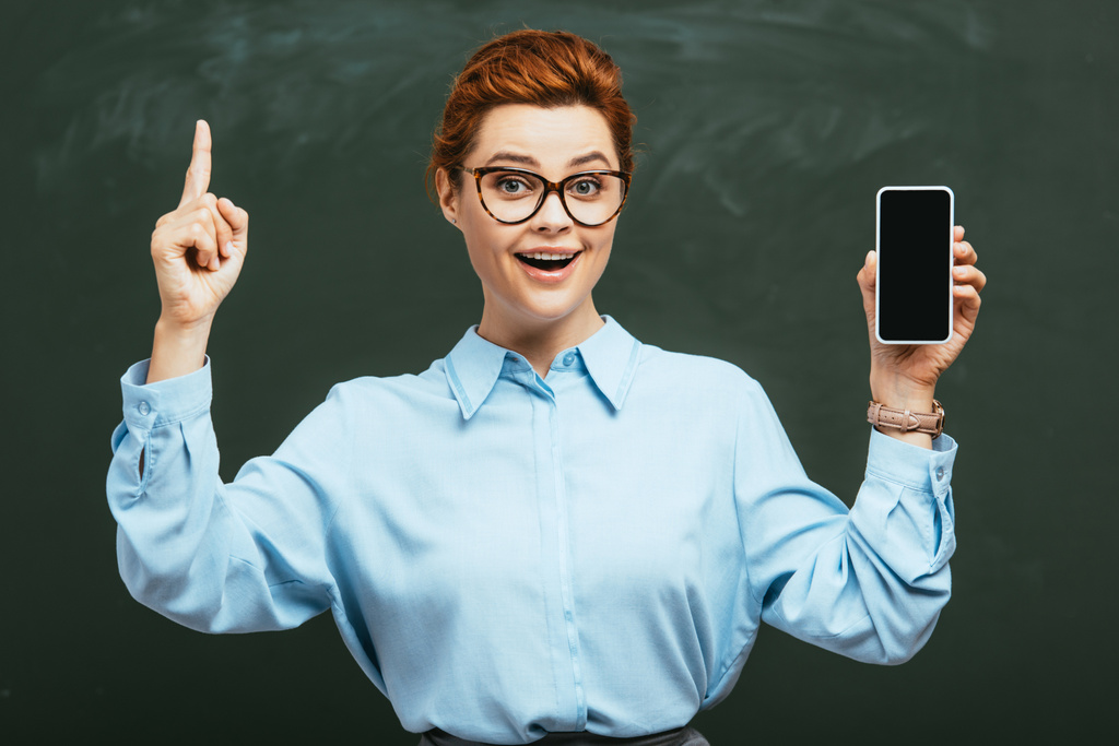 cheerful teacher in eyeglasses holding smartphone with blank screen near chalkboard    - Photo, Image