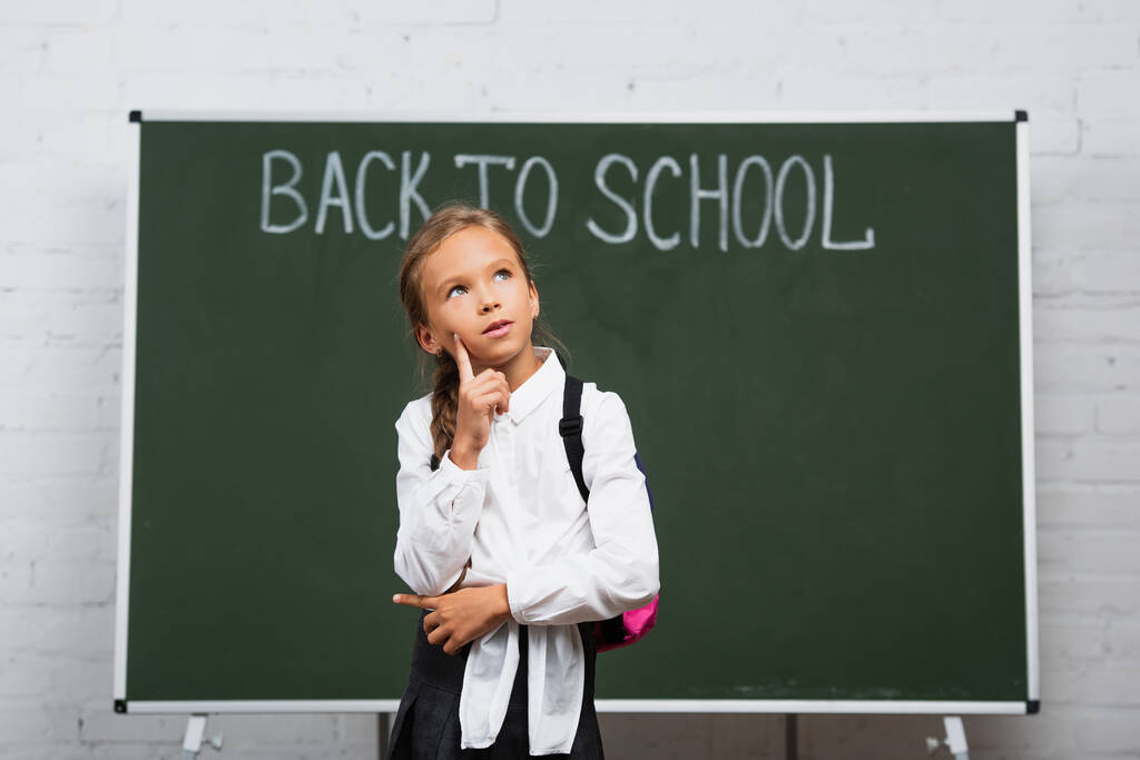 huomaavainen koulutyttö koskettaa kasvoja ja etsii lähellä liitutaulu takaisin kouluun kirjoitus  - Valokuva, kuva