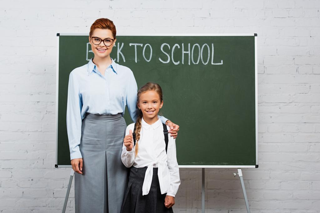 mosolygó tanár megérinti vállát boldog iskolás lány közelében táblán vissza az iskolai betű - Fotó, kép