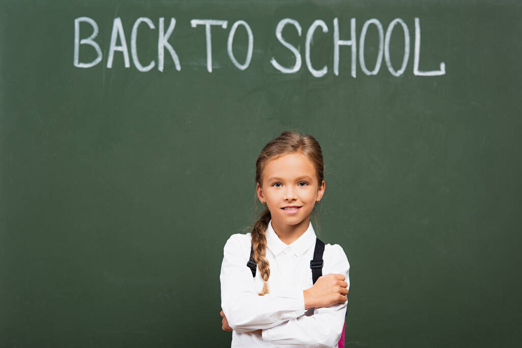 écolière souriante debout avec les bras croisés près du tableau noir avec inscription de retour à l'école - Photo, image