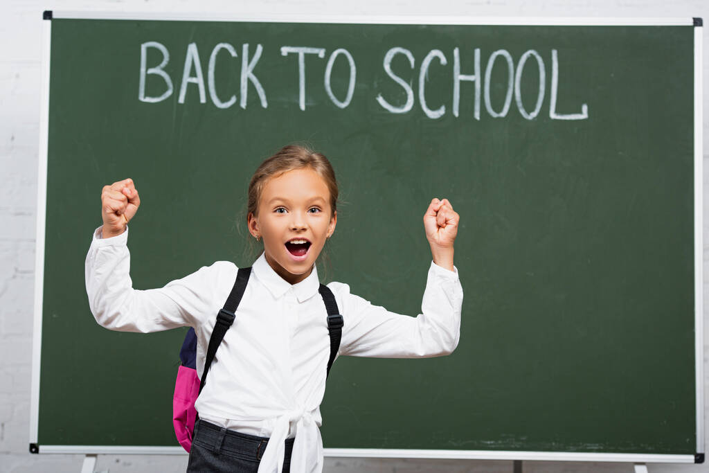 Aufgeregtes Schulmädchen zeigt Siegergeste neben Kreidetafel mit Schulschriftzug  - Foto, Bild