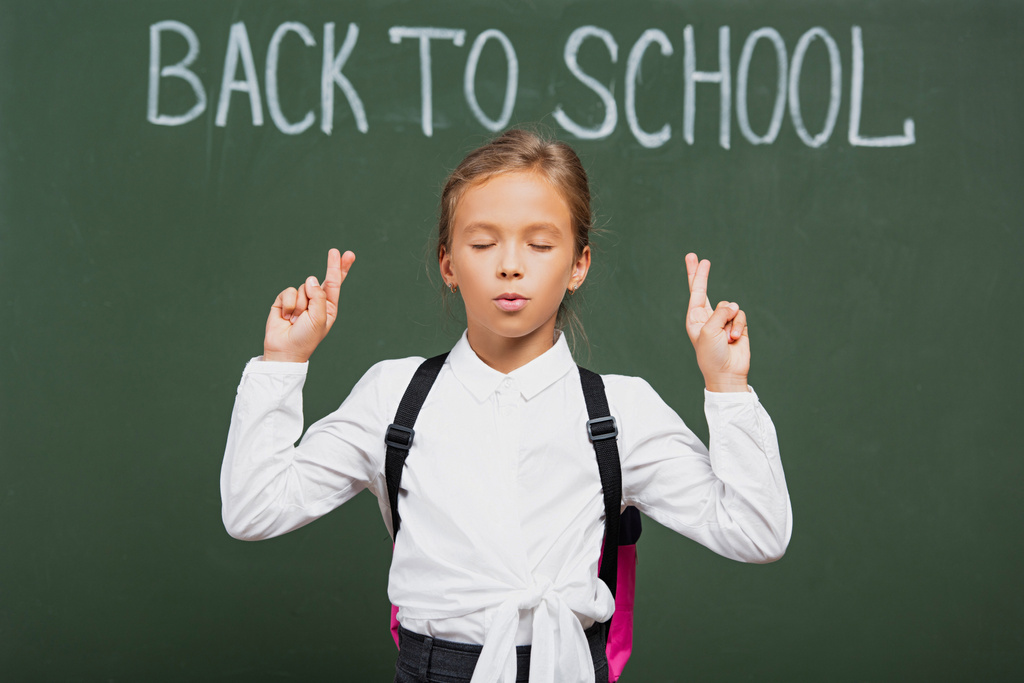schattig schoolmeisje met gesloten ogen houden gekruiste vingers in de buurt van terug naar school inscriptie op schoolbord - Foto, afbeelding
