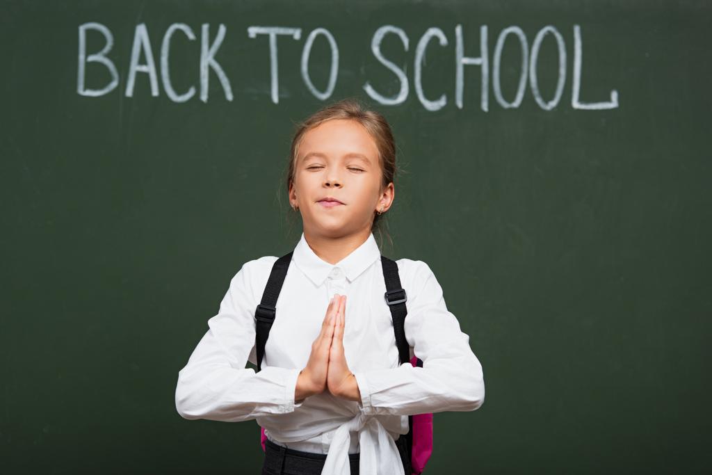 linda colegiala con los ojos cerrados sosteniendo las manos de oración cerca de nuevo a las letras de la escuela en pizarra
 - Foto, imagen