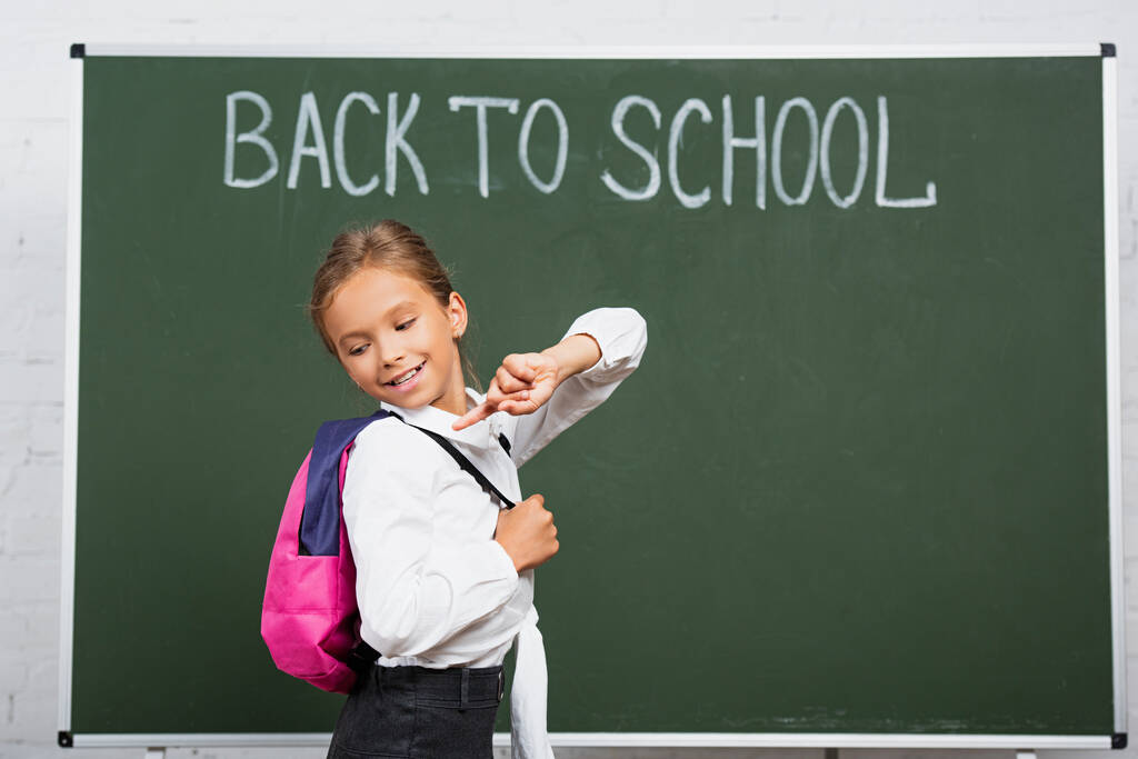 smiling schoolgirl pointing with finger at backpack near chalkboard with back to school inscription - Photo, Image