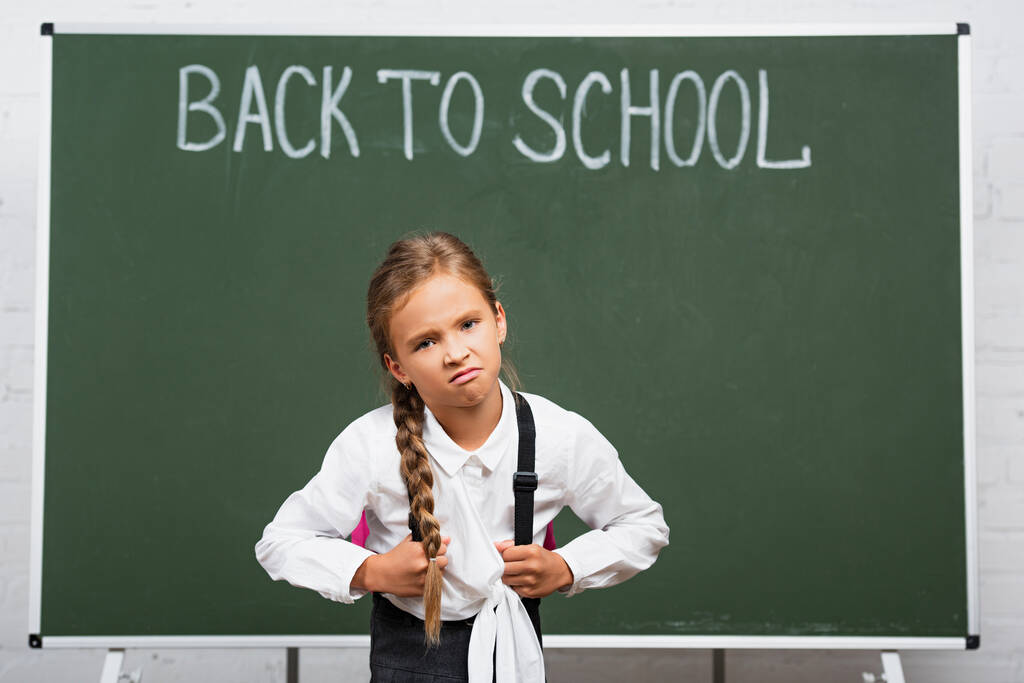 scolaretta dispiaciuta con zaino pesante vicino alla lavagna con lettere di ritorno a scuola  - Foto, immagini