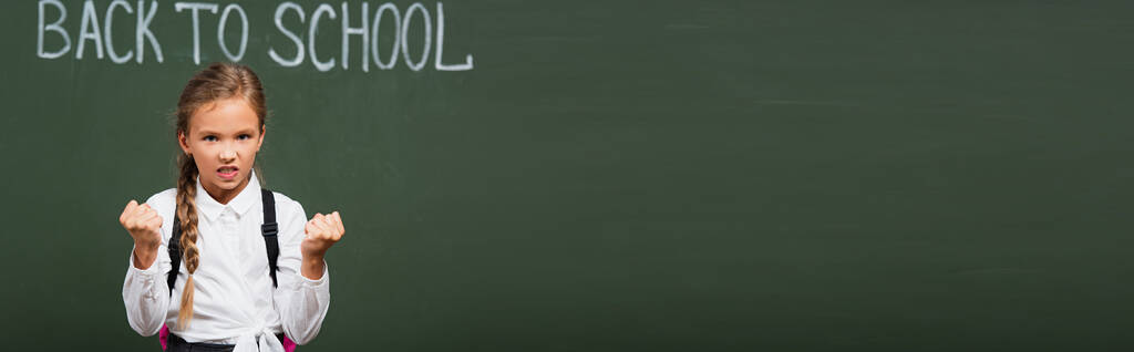 горизонтальное изображение разгневанной школьницы, показывающей сжатые кулаки возле доски с надписью - Фото, изображение