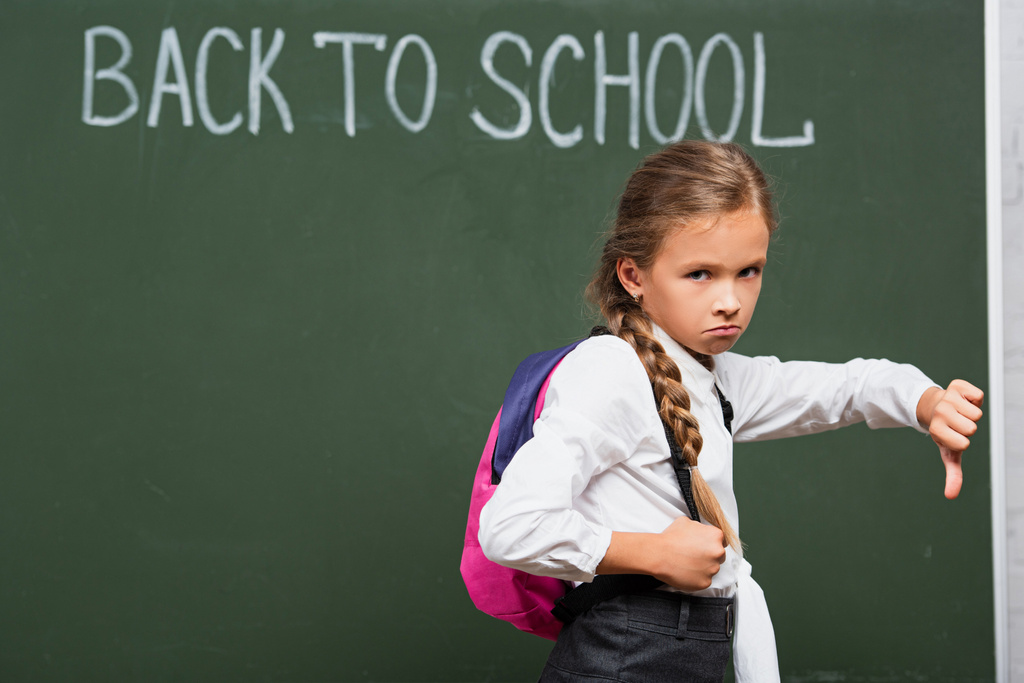 beledigd schoolmeisje tonen duim naar beneden en kijken naar camera in de buurt schoolbord met terug naar school belettering  - Foto, afbeelding