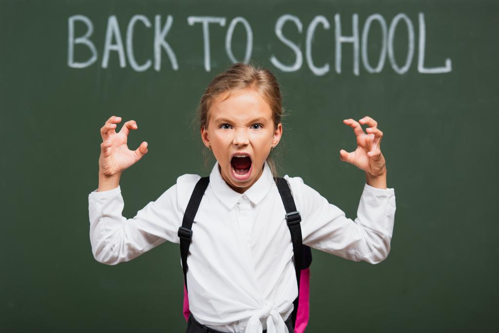 επιλεκτική εστίαση της θυμωμένος μαθήτρια δείχνει τρομακτικό χειρονομία κοντά chalkboard με πίσω στο σχολείο επιστολόχαρτα - Φωτογραφία, εικόνα