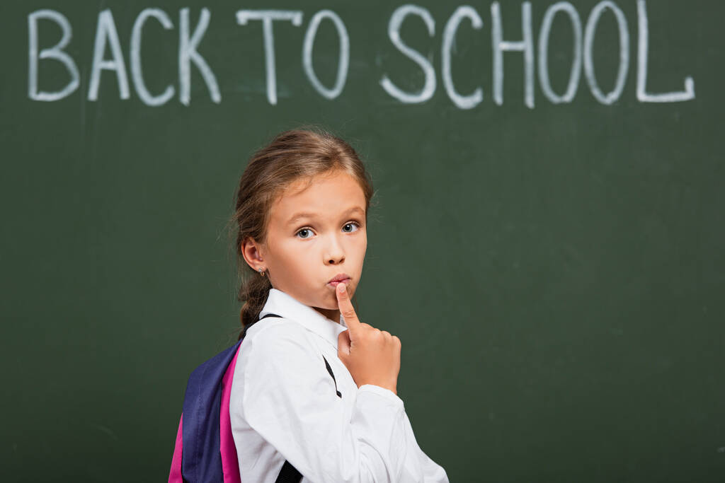 thoughtful schoolgirl touching lips and looking at camera near back to school inscription on chalkboard - Photo, Image