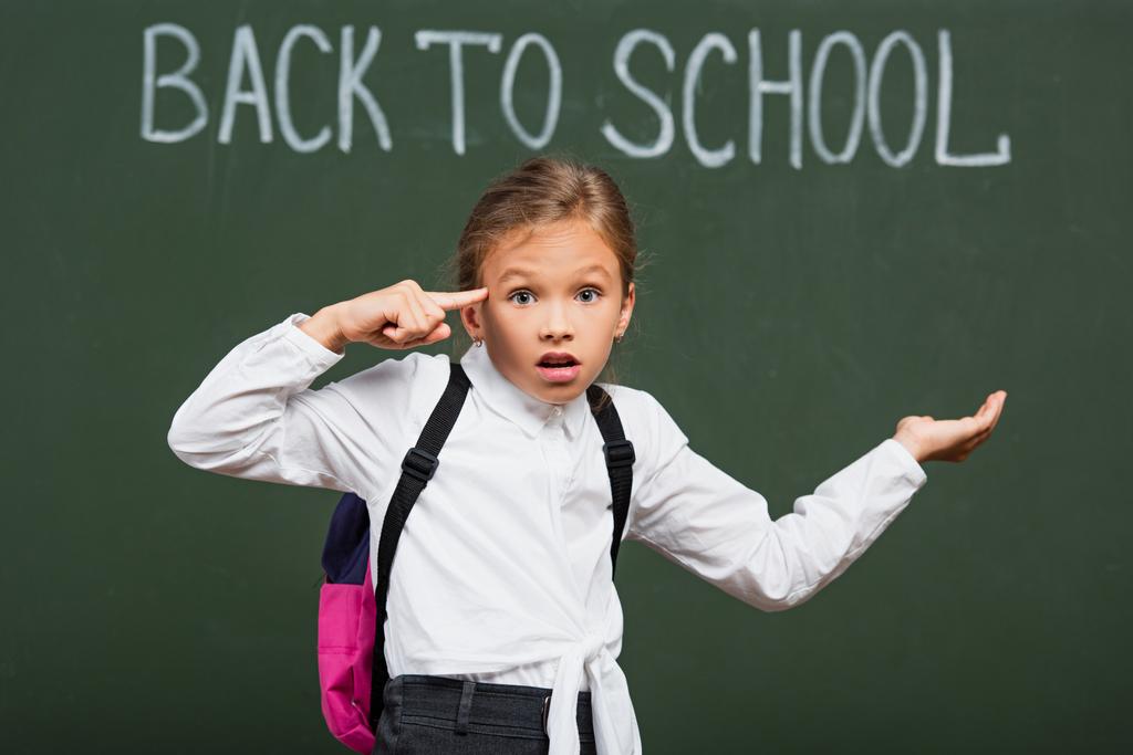 επιλεκτική εστίαση αποθαρρυμένη μαθήτρια δείχνει τρελό χειρονομία, ενώ στέκεται με ανοιχτό χέρι κοντά πίσω στο σχολείο επιγραφή στο μαυροπίνακα - Φωτογραφία, εικόνα