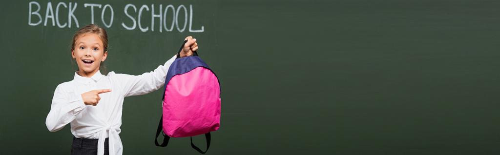 panoramatická úroda usměvavé školačky ukazuje na růžový batoh v blízkosti školní nápis na tabuli - Fotografie, Obrázek