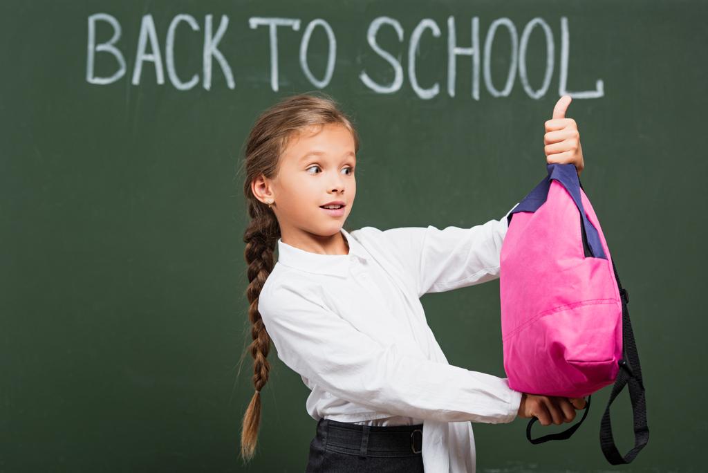 scolaretta sorpresa che tiene lo zaino rosa vicino alla lavagna con lettere di ritorno a scuola  - Foto, immagini