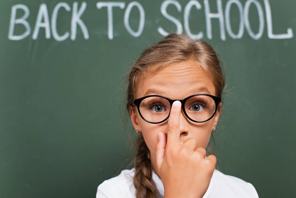 επιλεκτική εστίαση έκπληκτος μαθήτρια αγγίζοντας γυαλιά, ενώ κοιτάζοντας κάμερα κοντά πίσω στο σχολείο γράμματα σε πίνακα - Φωτογραφία, εικόνα