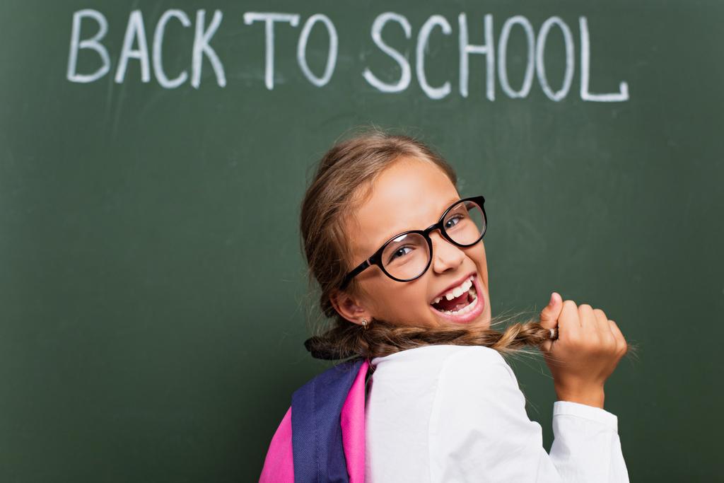избирательный фокус возбужденной школьницы в очках, касающихся волос и смеющихся возле доски со школьными надписями - Фото, изображение