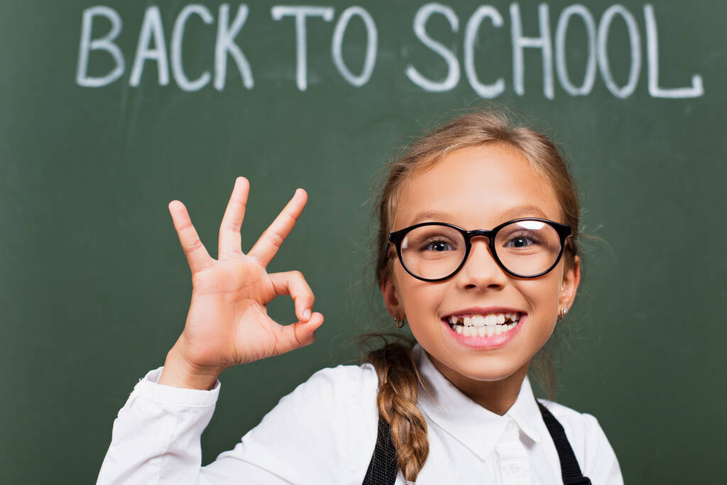 избирательный фокус возбужденной школьницы в очках, показывающей большой палец вверх рядом с доской с текстом из школы - Фото, изображение